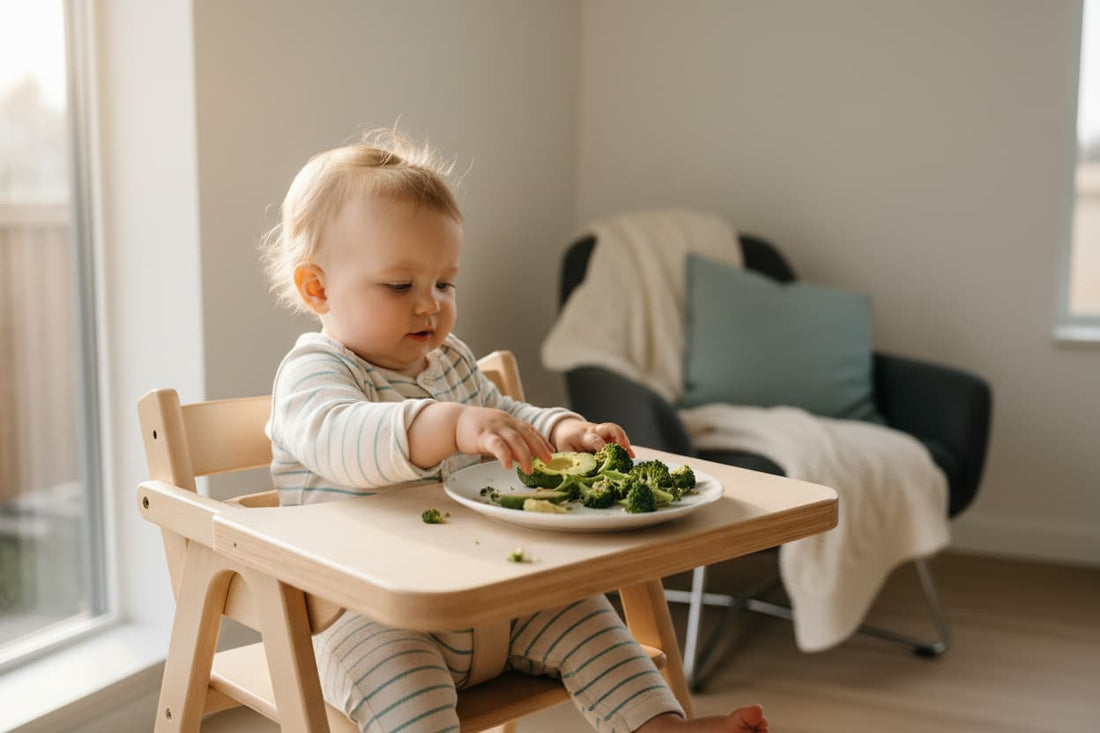 Baby sitting in a high chair reaching for soft finger foods including broccoli and avocado slices — baby-led weaning first foods